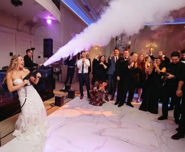 feestelijke uitbarsting van bruid met Co2 kanon Een bruid in bruidsjurk vuurt Co2 rookkanon af op dansvloer terwijl gasten juichen. Op de achtergrond draait een bruiloft dj achter de dj booth.