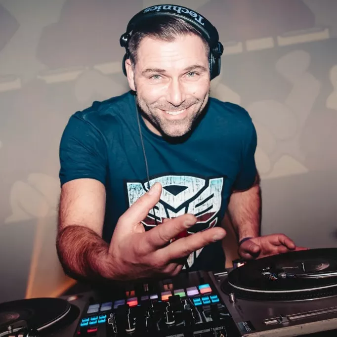 DJ DNS tijdens een energieke set achter de draaitafels DJ DNS (Dennis Bruijn) draait glimlachend achter de draaitafels met koptelefoon op en een shirt met het Transformers-logo, terwijl hij het publiek aanspreekt in een energieke clubsfeer.